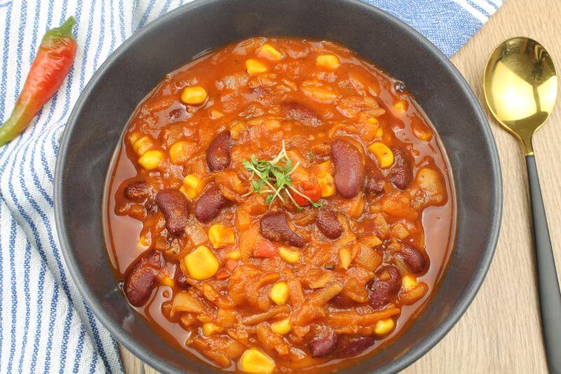 Ein Teller mit Chilli sin Carne ohne Beilagen, rechts daneben ein Löffel, links eine Chillischote